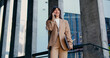 © serg - Confident businesswoman in a formal suit holding a laptop and talking on the phone while walking outdoors. Modern glass office building in the background. Business communication, important call