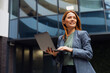 © Novak - Successful business woman in a formal suit standing and using a laptop in front of a modern glass office building in the city.