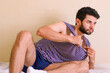 © Cristiano Costa - Bearded man lying in bed getting up in the morning. Handsome young man with a fit body getting out of bed, waking up. Wearing a blue shirt and blue underwear in the bedroom. Legs and arms visible.