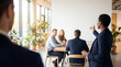 © Natália  - Businesswoman speaking to colleagues in modern open plan office. Manager addressing team at wooden table for leadership meeting, employee onboarding and company workshop.