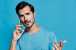 © SHOTPRIME STUDIO - Concerned young man talking on mobile phone, wearing blue t-shirt, looking thoughtfully at camera against vibrant blue background. Communication lifestyle concept.