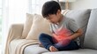 © Ohana - Asian boy sitting on sofa suffering from stomach ache with red pained area effect on abdomen