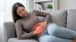 © Ohana - Young Asian woman sitting on sofa suffering from stomach ache and menstrual pain with red glow effect on abdomen
