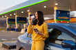 © Home-stock - Lady standing by her car at parking lot near gas station while sipping coffee and using cellphone, taking break during road trip and checking route