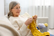 © New Africa - Senior woman knitting with needles on sofa at home