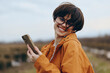 © SHOTPRIME STUDIO - Smiling woman lifestyle wearing glasses and orange jacket holds smartphone outdoors in natural environment. Happy young female enjoys technology and casual fashion concept.