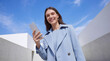 © AAUB - Low angle view of a smiling young woman in a blue coat holding a smartphone under a clear blue sky. Concept for digital connectivity, mobile lifestyle, modern business, and urban life.