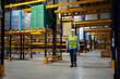 © Videophilia - A person in a safety vest and helmet walks through an empty warehouse. The space has high racks filled with boxes and products in rows. Bright lights illuminate the area.