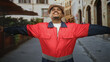 © Krakenimages.com - Man construction worker wearing a hard hat and red safety jacket, beard visible, arms outstretched with open palms while standing on a cobblestone street, smiling; pride duty.