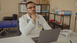 © Krakenimages.com - Young hispanic man in white coat leans on desk with hand on chin in clinic office among medical shelves; thinking doubt uncertainty.