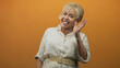 © Krakenimages.com - Senior woman waves hand and smiles in studio with orange backdrop and visible jewelry; welcoming joy.