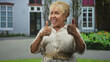 © Krakenimages.com - Senior woman shows two thumbs up gesture and smiling face in front of building with garden and windows; joy confidence.