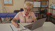 © Krakenimages.com - Senior woman physiotherapist with stethoscope writing on clipboard at clinic and consulting a laptop; calm concentration.