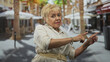 © Krakenimages.com - Senior hispanic woman pointing with both hands and extended fingers in a sunny street cafe with palm trees and umbrellas, directing attention to the right; confidence.