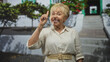 © Krakenimages.com - Senior hispanic woman holds up peace sign with hand near face, beige linen shirt and belt in front of building entrance; joy confidence.