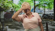 © Krakenimages.com - Senior hispanic woman covering eyes with hand while smiling and waving in a city street with bicycles and trees visible; joy playfulness.