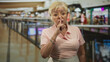 © Krakenimages.com - Woman senior hispanic with finger to lips shushing gesture in a busy building corridor near railing and shops, wearing glasses and bracelets; quiet warning.