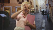 © Krakenimages.com - Woman senior smiling, wearing glasses and bracelet, holding pink clipboard close to chest and making ok sign with hand in a street; cheerful travel confidence.