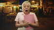 © Krakenimages.com - Senior woman clutching pink clipboard with open mouth gesture on a night street; surprise curiosity.