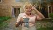 © Krakenimages.com - Woman holding stovetop coffee maker and pointing finger to temple in front of rustic stone building with courtyard path; contemplation.