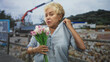 © Krakenimages.com - Senior hispanic woman holding pink tulips touches neck on a street with visible construction crane and stone wall; quiet contemplation.