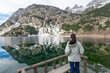 © Fominayaphoto - Woman standing on wooden viewpoint enjoying a calm mountain lake with perfect reflection in Panticosa, Pyrenees, Spain. Winter landscape with snow and peaceful atmosphere.
