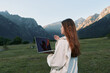 © SHOTPRIME STUDIO - A focused outdoor professional stands with a laptop in a mountain valley. She wears light outerwear, gazing ahead with confidence, creating a calm, productive, and inspirational work scene.