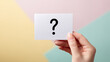 © photo for everything - A feminine hand gently holds a white card featuring a bold black question mark symbol, set against a vibrant, pastel geometric background