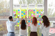 © Pixel-Shot - Group of young people discussing business plan at Kanban board on window in office, back view