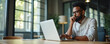 © Vadym - Black man in glasses typing on laptop computer. He wears light shirt, sits at table in office. Man looks thoughtful, holding hand near mouth, thinking about work.
