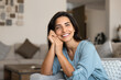 © fizkes - Calm Latina woman sitting comfortable on cozy sofa, looks at camera with contented smile, enjoying weekend rest in fashionable living room, spend weekend, daydreaming indoors. Homeowner lady portrait