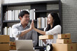© AU USAnakul - Asian couple entrepreneurs high fiving each other in home office surrounded by packages and laptop, smiling and celebrating success, e commerce business growth and teamwork concept