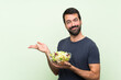 © luismolinero - Young handsome man with salad over isolated green wall extending hands to the side for inviting to come