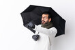 © luismolinero - Caucasian handsome man with beard holding an umbrella over isolated white wall pointing away