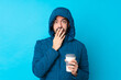 © luismolinero - Man wearing winter jacket and holding a takeaway coffee over isolated blue background surprised and shocked while looking right