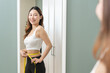 © KMPZZZ - Weight lose, loss concept. Slim body asian young woman hand use tape measuring around waistline in fit sports. Girl looking reflect in mirror at home. Healthy nutrition, fitness for wellbeing beauty.