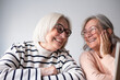 © luciano - Two smiling senior women with white hair and glasses sitting at a wooden table at home, looking at a laptop together. They appear relaxed and happy while browsing online, with papers and a coffee mug