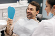 © Home-stock - Happy male patient smiling while looking in mirror during dental appointment at clinic, being satisfied with result of procedure