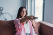 © Daniel - Young woman lounges on a couch in a bright living room, smiling and holding a TV remote as she relaxes and watches television, enjoying leisure time at home.