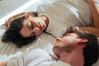 © Daniel - A young couple lies facing each other on a white bed, exchanging warm smiles and gentle eye contact, conveying intimacy, comfort and peaceful morning togetherness in a bright bedroom.