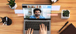 © Prostock-studio - A woman is seen working at her office desk, typing on a laptop while engaged in an online call. The screen shows a smiling man with several colleagues visible in a video meeting.