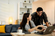 © Thitiporn - Young couple working together on laptop at home, man pointing at screen helping woman study and write notes. Remote lifestyle, teamwork, productivity, online learning, cozy home interior