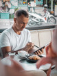 © Aboltin - Man using tablet in cafe while finishing meal, working remotely. Digital habits, online business and flexible lifestyle concept.