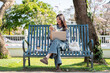 © bongkarn - Pretty asian woman looking at phone over laptop while sitting cross legged on bench in garden park.