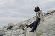 © SHOTPRIME STUDIO - Young woman sits on rocky shore wearing casual clothes, looking away and smiling. Overcast sky creates calm atmosphere by the sea. Concept of relaxation and nature escape.
