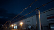 © photo for everything - Barbed wire fence with chain link against a dark blue evening sky, warm lights blur in background, creating a mood of security and exclusion