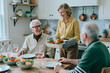 © AnnaStills - Senior woman serving roasted chicken to senior friends during home dinner, celebrating togetherness and active retirement