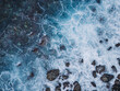 © luciano - Aerial top-down view of turquoise ocean waves crashing against a rugged rocky shoreline. The dynamic movement of white sea foam contrasts with dark volcanic stones, creating a dramatic natural texture