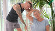 © luciano - Active elderly woman exercising inside home with a personal trainer stretching exercises for shoulders and arms in a modern living room. Healthy  lifestyle, strength training, and wellness concept