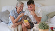 © luciano - A 70-year-old woman and a young 20-year-old grandson look at the boy's cell phone together while sitting on the couch at home. Friendship and affection in a multigenerational family